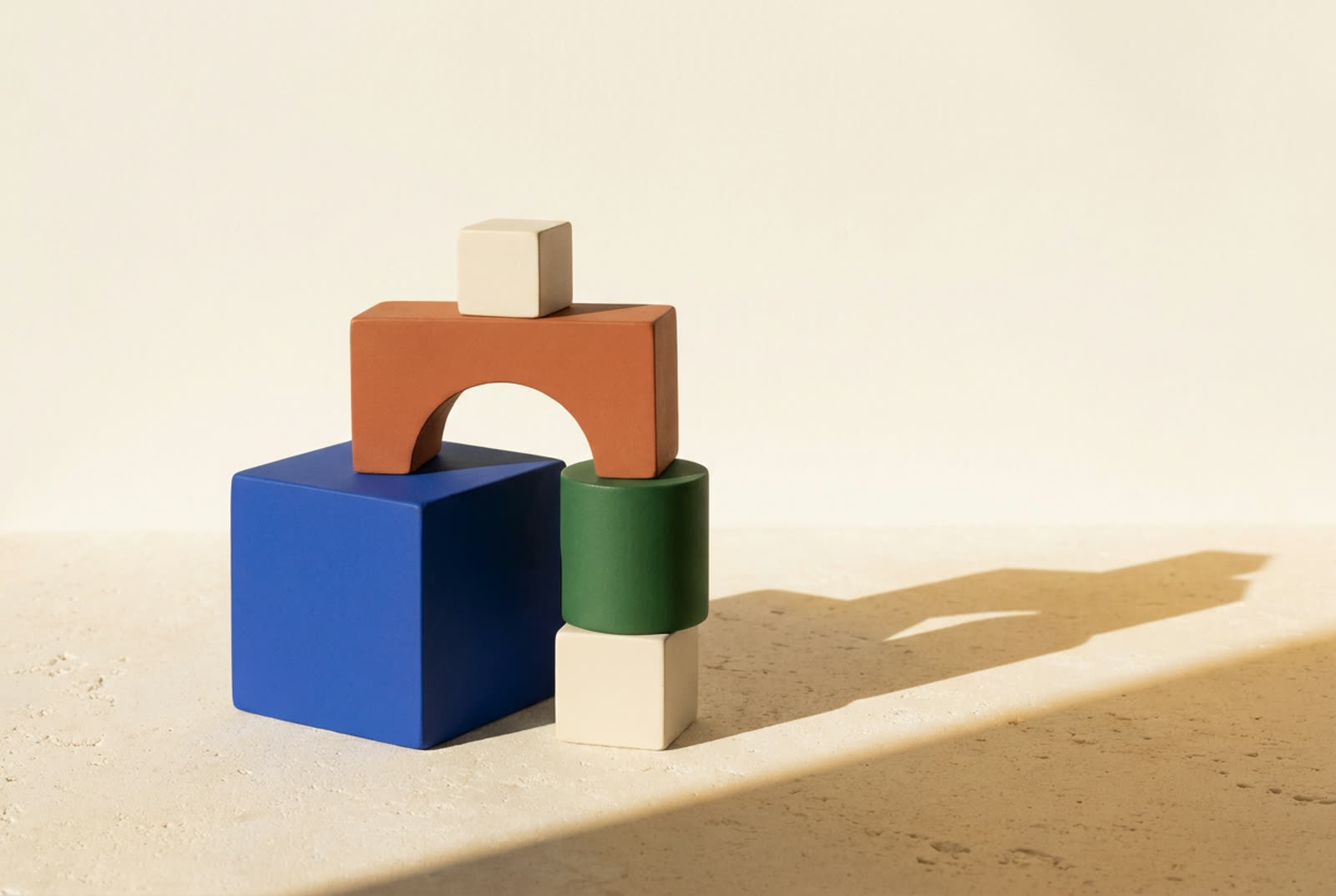 Matte geometric blocks — an arch, a cube and a cylinder — in cobalt, terracotta and green, casting long shadows on warm limestone